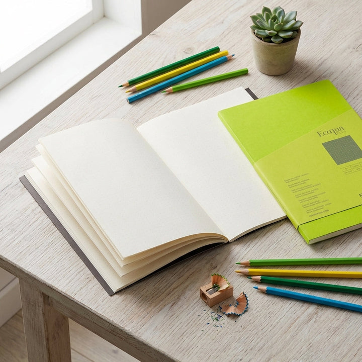 Creative flat lay of open Fabriano Ecoqua Plus Block with colorful pencils and plant, emphasizing vibrant lime cover and paper texture.